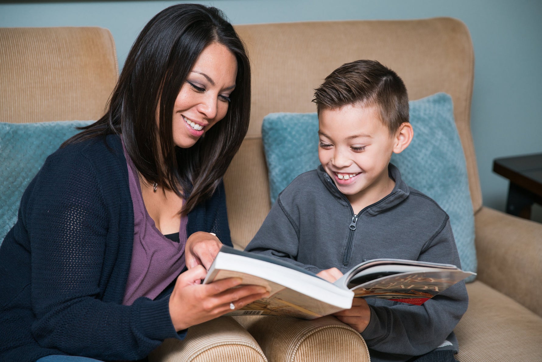 A heartwarming photo of a woman and a young boy sitting together on a couch, smiling as they read a large book together.